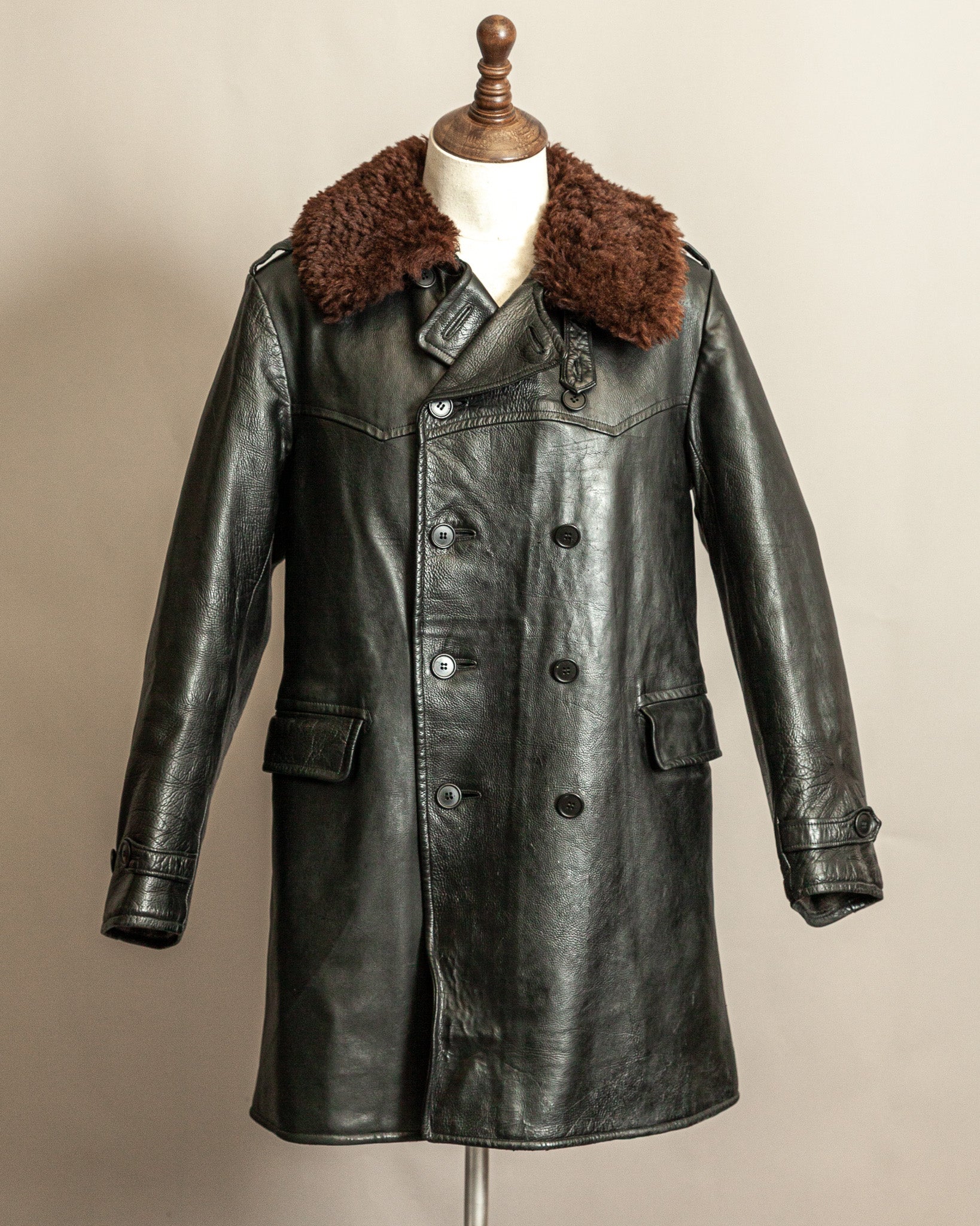 Vintage Swedish WW2 1940s Black Goatskin leather long army trench coat with real wool collar on a mannequin against a beige background, the top button is undone and the lapels are showing slightly open, the jacket is displaying grained leather