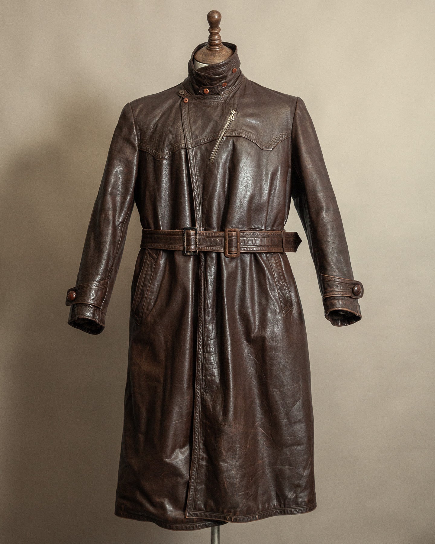 Vintage brown German officer-style horsehide leather trench coat with belted waist, throat latch, and long silhouette, displayed on a mannequin against a neutral background.