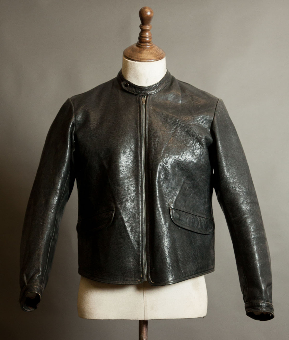Front of a vintage European black goatskin motorcycle jacket with band collar, grey Riri front zip and curved flap pockets, showing natural patina