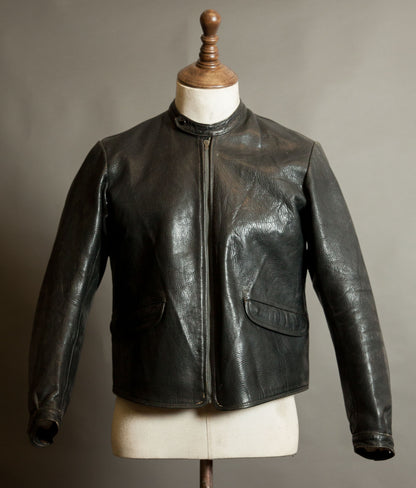 Front of a vintage European black goatskin motorcycle jacket with band collar, grey Riri front zip and curved flap pockets, showing natural patina
